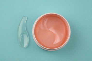Plastik bir kabın içindeki göz bantları, üst manzara. Göz çevresi için peptit içeren hidrojel yamalar. Mavi arka plandaki kavanozda plastik spatula bulunan pembe jel yamalar.. 