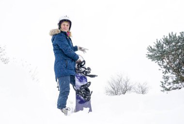 Kız elinde bir snowboard tutuyor, dağ ceketi ve kask giyiyor.