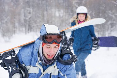 Snowboard kıyafetleri içinde bir adam ve bir kız kayak yamacı boyunca yürüyorlar. Bir snowboard tutuyorlar. Kış tatili dağlarda.