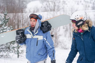 Snowboard kıyafetleri içinde bir adam ve bir kız kayak yamacı boyunca yürüyorlar. Bir snowboard tutuyorlar. Kış tatili dağlarda.