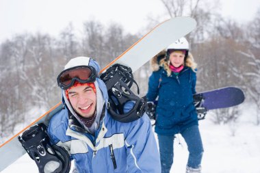 Snowboard kıyafetleri içinde bir adam ve bir kız kayak yamacı boyunca yürüyorlar. Bir snowboard tutuyorlar. Kış tatili dağlarda.