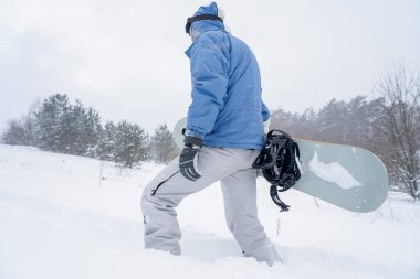 Snowboard yapan adam karlı bir yamaca çıkıyor. Elinde bir snowboard tutuyor.