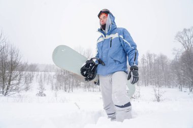 Snowboard yapan adam karlı bir yamaca çıkıyor. Elinde bir snowboard tutuyor.