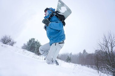 Snowboard yapan adam karlı bir yamaca çıkıyor. Elinde bir snowboard tutuyor.