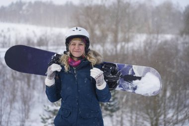 Kız elinde bir snowboard tutuyor, dağ ceketi ve kask giyiyor.