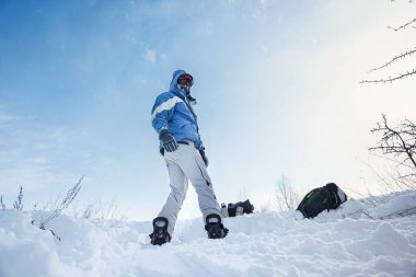 Snowboardcu elinde kayak ekipmanıyla dağın yamaçlarında duruyor.