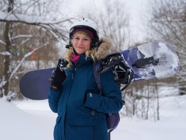Kız elinde bir snowboard tutuyor, dağ ceketi ve kask giyiyor.