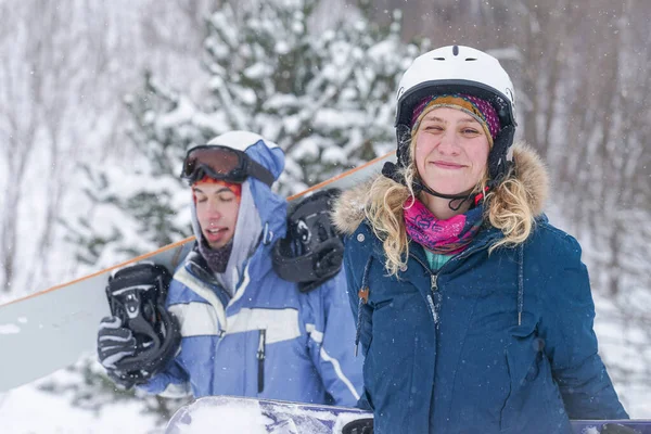 Snowboard kıyafetleri içinde bir adam ve bir kız kayak yamacı boyunca yürüyorlar. Bir snowboard tutuyorlar. Kış tatili dağlarda.