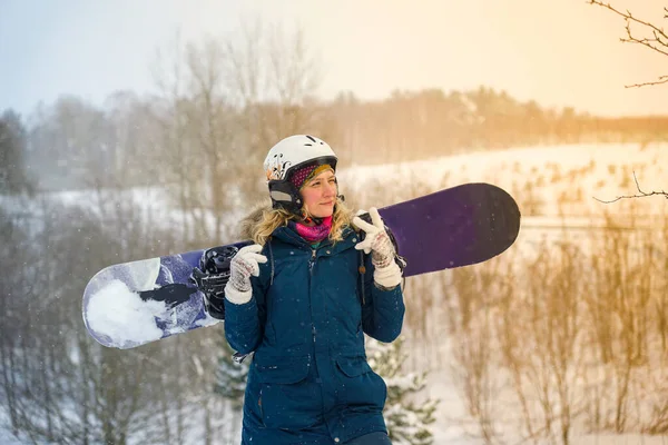 Kız elinde bir snowboard tutuyor, dağ ceketi ve kask giyiyor.