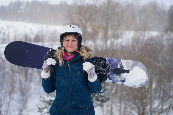 Kız elinde bir snowboard tutuyor, dağ ceketi ve kask giyiyor.