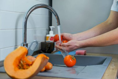 Aşçı musluktan akan suyun altında taze sebzeleri yıkar. Elinde iki tane olgun kırmızı domates tutuyor.