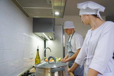 Two chefs in the restaurant kitchen prepare food. The chef is dressed in a white tunic and a chef's hat.	