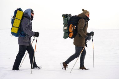 İki adam kış gezisi sırasında gevşek karda yürür. Büyük sırt çantaları, kalın ceketler taşıyorlar. Ellerinde yürüyüş sopaları tutarlar..