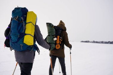 İki adam kış gezisi sırasında gevşek karda yürür. Büyük sırt çantaları, kalın ceketler taşıyorlar. Ellerinde yürüyüş sopaları tutarlar..