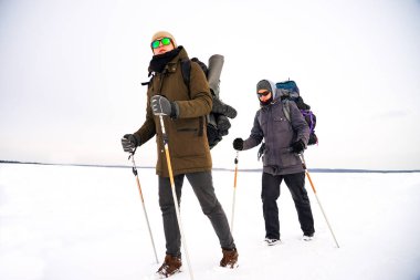 İki adam kış gezisi sırasında gevşek karda yürür. Büyük sırt çantaları, kalın ceketler taşıyorlar. Ellerinde yürüyüş sopaları tutarlar..