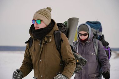 İki adam kış gezisi sırasında gevşek karda yürür. Büyük sırt çantaları, kalın ceketler taşıyorlar. Ellerinde yürüyüş sopaları tutarlar..
