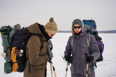 İki adam kış gezisi sırasında gevşek karda yürür. Büyük sırt çantaları, kalın ceketler taşıyorlar. Ellerinde yürüyüş sopaları tutarlar..