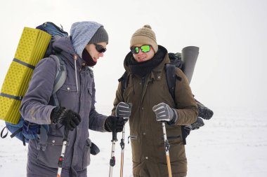 İki adam kış gezisi sırasında gevşek karda yürür. Büyük sırt çantaları, kalın ceketler taşıyorlar. Ellerinde yürüyüş sopaları tutarlar..