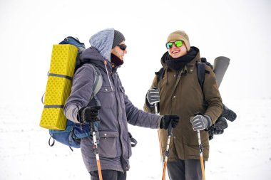 İki adam kış gezisi sırasında gevşek karda yürür. Büyük sırt çantaları, kalın ceketler taşıyorlar. Ellerinde yürüyüş sopaları tutarlar..
