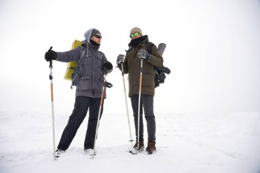 İki adam kış gezisi sırasında gevşek karda yürür. Büyük sırt çantaları, kalın ceketler taşıyorlar. Ellerinde yürüyüş sopaları tutarlar..
