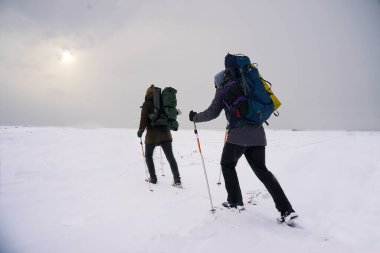 Adamlar kış gezisi sırasında konuşuyorlar. Onlar kalın ceketler, şapkalar ve büyük sırt çantaları giyiyorlar. Karda durup yürüyüş sopalarını tutarlar.