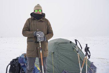 Şapkalı ve gözlüklü bir adam elinde karlı bir vadinin arka planına değnek tutuyor.