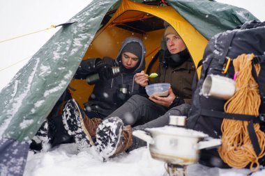 Çocuklar bir çadırda oturuyor, kış yürüyüşü sırasında yemek hazırlıyorlar. Karda çadırın yanında ekipmanları yatıyor. İki adam çadırda yemek yiyor. Keşif sırasında kışın turistlere yemek servisi.