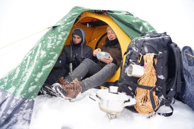 Çocuklar bir çadırda oturuyor, kış yürüyüşü sırasında yemek hazırlıyorlar. Karda çadırın yanında ekipmanları yatıyor. İki adam çadırda yemek yiyor. Keşif sırasında kışın turistlere yemek servisi.