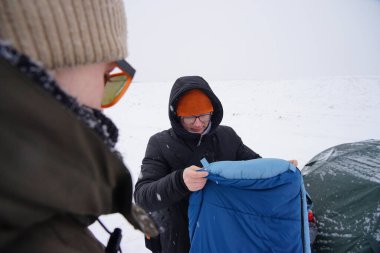 Adamın elinde uyku tulumu var. Kışın çadırın yanında durur.