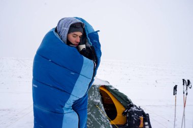 Uyku tulumunda bir adam çadırın yanındaki karda duruyor ve kahve içiyor.