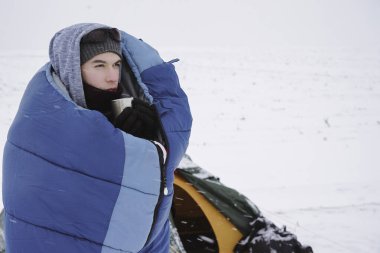 Uyku tulumunda bir adam çadırın yanındaki karda duruyor ve kahve içiyor.