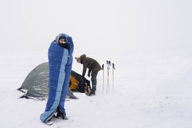 Uyku tulumunda bir adam çadırın yanındaki karda duruyor ve kahve içiyor.