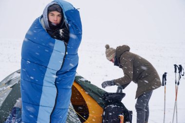 Uyku tulumunda bir adam çadırın yanındaki karda duruyor ve kahve içiyor.