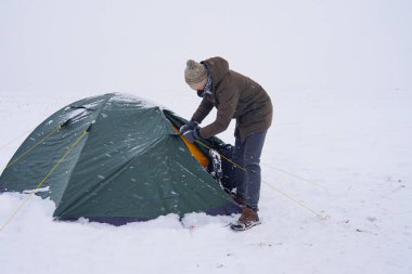 Bir turist karda çadırın yanında oturur, bir kış fırtınası sırasında çadırın sabitlenmesi için sırt çantasından bir kordon çıkarır.