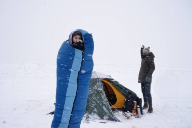 Uyku tulumunda bir adam çadırın yanındaki karda duruyor ve kahve içiyor.