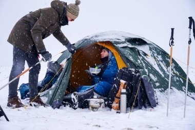 Çocuklar bir çadırda oturuyorlar, kış tatili boyunca yemek hazırlıyorlar. Karda çadırın yanında ekipmanları var.