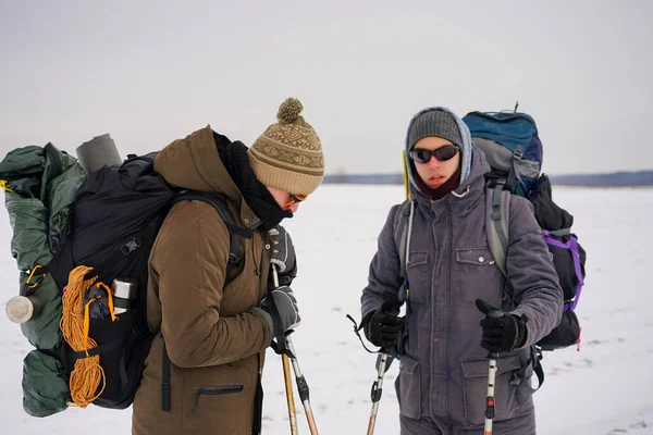 İki adam kış gezisi sırasında gevşek karda yürür. Büyük sırt çantaları, kalın ceketler taşıyorlar. Ellerinde yürüyüş sopaları tutarlar..