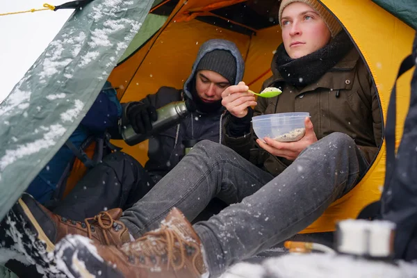 Çocuklar bir çadırda oturuyor, kış yürüyüşü sırasında yemek hazırlıyorlar. Karda çadırın yanında ekipmanları yatıyor. İki adam çadırda yemek yiyor. Keşif sırasında kışın turistlere yemek servisi.
