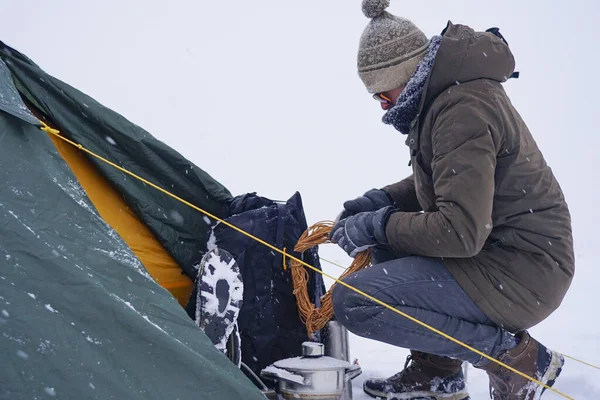 Bir turist karda çadırın yanında oturur, bir kış fırtınası sırasında çadırın sabitlenmesi için sırt çantasından bir kordon çıkarır.