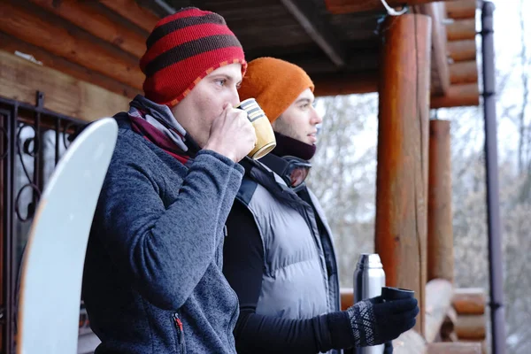 Friends chat after skiing. They stand on the balcony of a wooden house in winter clothes, drink tea and relax. Porter of two guys in a ski resort in the alps