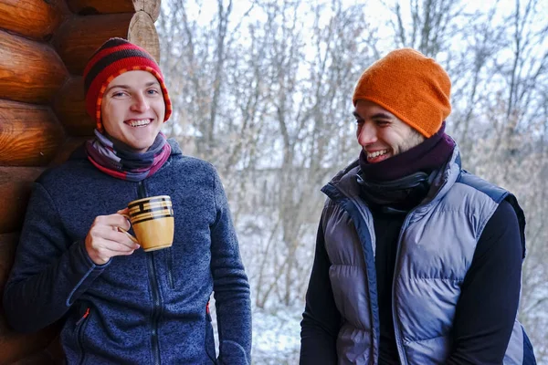Two guys chat after skiing. They stand on the balcony of a wooden house in winter clothes, drink tea and relax. Porter of two guys in a ski resort in the alps