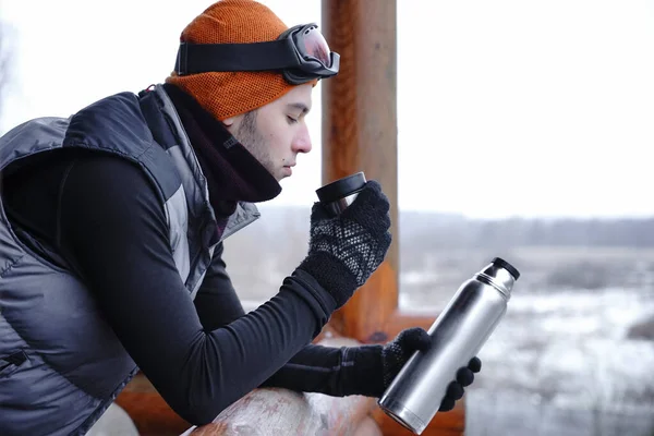 A guy in an orange hat in winter drinks tea from a thermos during a winter vacation in the mountains. He stands on the balcony of a wooden house. Portrait of a guy with a cup in his hands at a ski resort