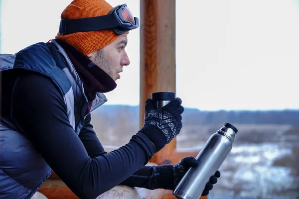 A guy in an orange hat in winter drinks tea from a thermos during a winter vacation in the mountains. He stands on the balcony of a wooden house. Portrait of a guy with a cup in his hands at a ski resort