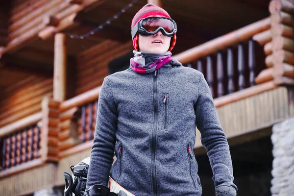 A guy in ski goggles holds a snowboard in his hand near the resort house in the ski base. Portrait