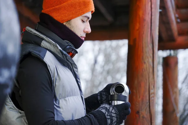 The guy stand on the balcony of a wooden house in winter clothes, drink tea and relax. Porter of two guys in a ski resort in the alps. Man pouring tea from a thermos