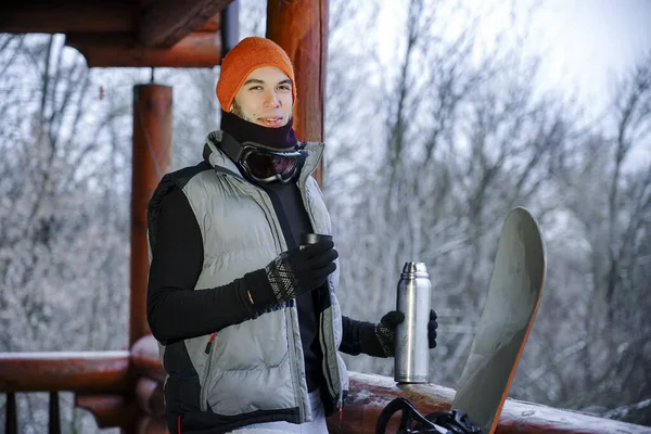 The guy stand on the balcony of a wooden house in winter clothes, drink tea and relax. Porter of two guys in a ski resort in the alps. Man pouring tea from a thermos