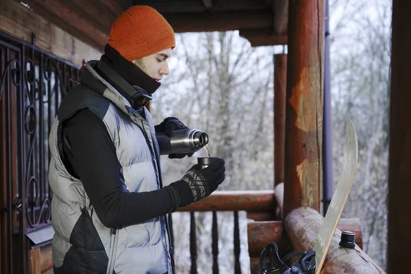 The guy stand on the balcony of a wooden house in winter clothes, drink tea and relax. Porter of two guys in a ski resort in the alps. Man pouring tea from a thermos