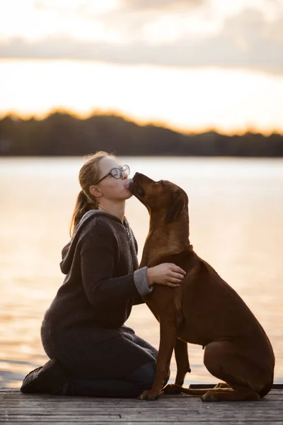 Flicka och hund kramas i solnedgång på vatten bakgrund — Stockfoto