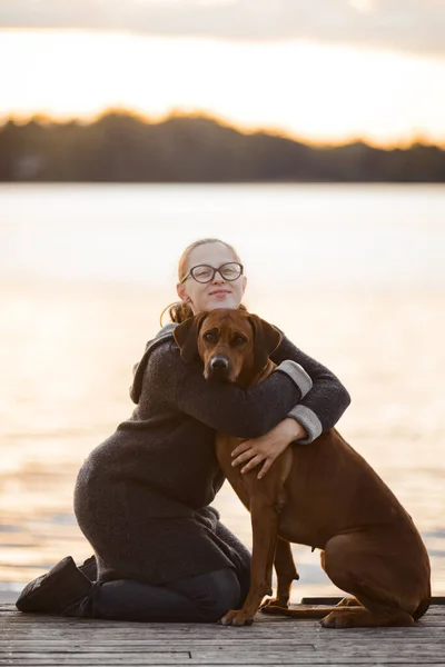 Girl and dog hugging in sunset on water background — стоковое фото