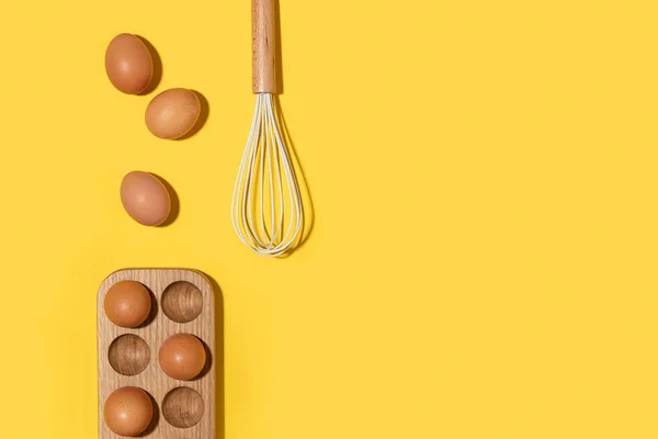Eggs in wooden egg box and whisk on yellow background. Flat lay Top view Copy space
